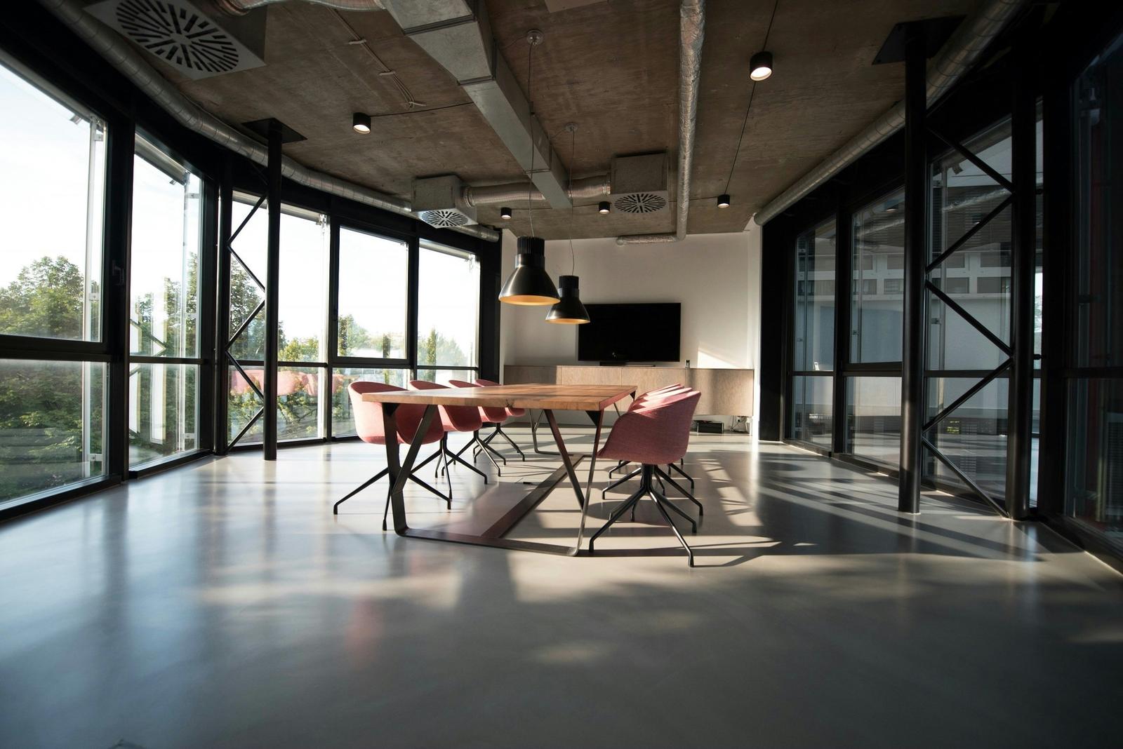 An empty meeting room with a long wooden table, pink chairs, and floor-to-ceiling windows.
